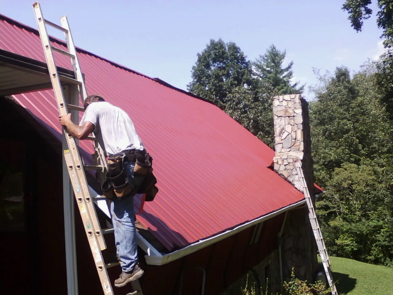 Roofer on a ladder beside a red metal roof and stone chimney — Soffit & Fascia Repair by Roofing Brothers in Winona Lake